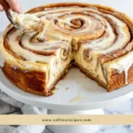 Whole cinnamon roll honeybun cheesecake with glazed top and a slice being removed
